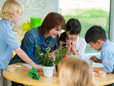 A group of young school children crowd around a table, while the teacher sitting at the table, talks to them while pointing at a piece of paper