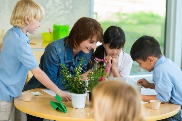 A group of young school children crowd around a table, while the teacher sitting at the table, talks to them while pointing at a piece of paper