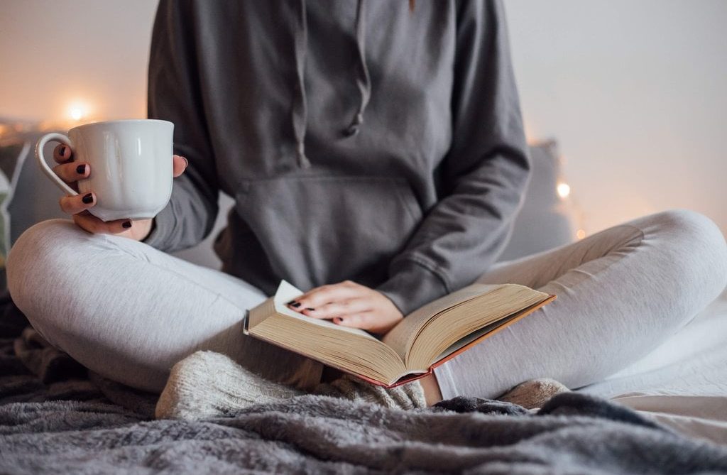 Someone sits crossed legged on a soft surface, holding a cup of tea/coffee while reading a book
