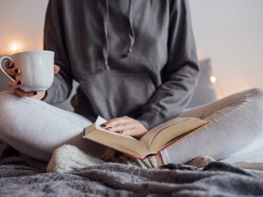 Someone sits crossed legged on a soft surface, holding a cup of tea/coffee while reading a book