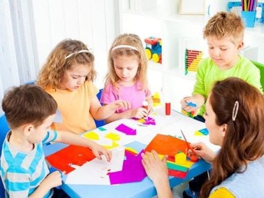 A group of young children sit around an arts & crafts table. Each child is dressed in bright colours & shows fascination in cutting shapes out of paper.