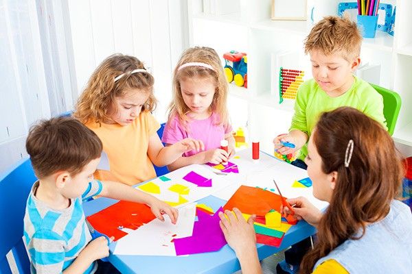A group of young children sit around an arts & crafts table. Each child is dressed in bright colours & shows fascination in cutting shapes out of paper.