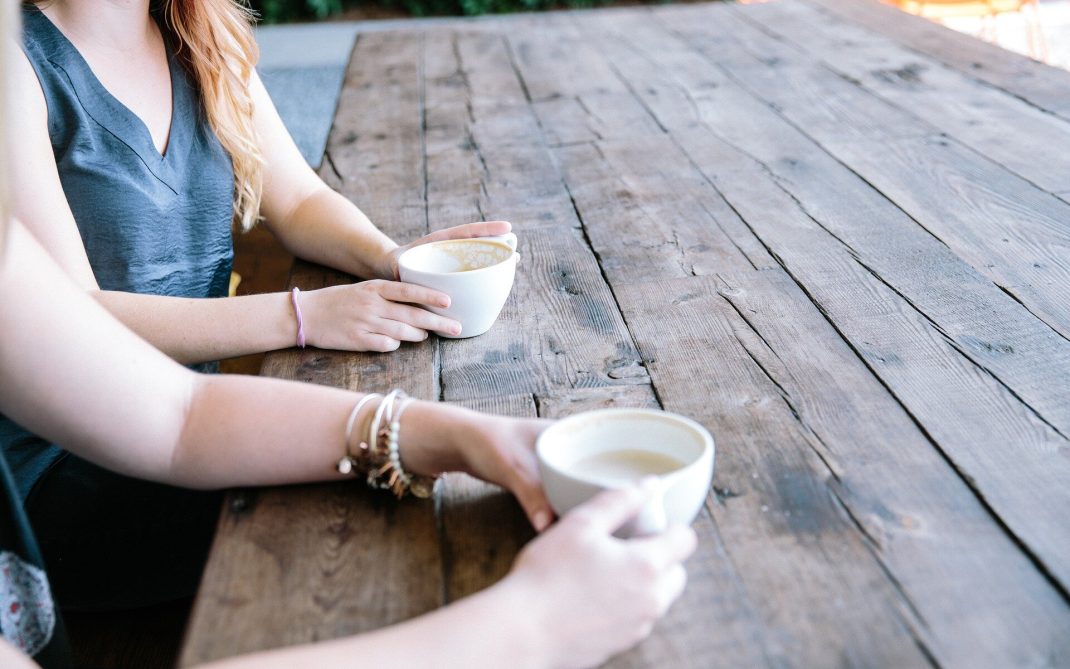 Two people sit at a rustic wooden table outdoors, each holding a white mug filled with a hot beverage. Only their arms and hands are visible, and the table stretches into the background.