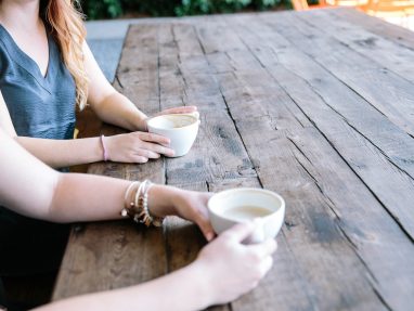 Two people sit at a rustic wooden table outdoors, each holding a white mug filled with a hot beverage. Only their arms and hands are visible, and the table stretches into the background.