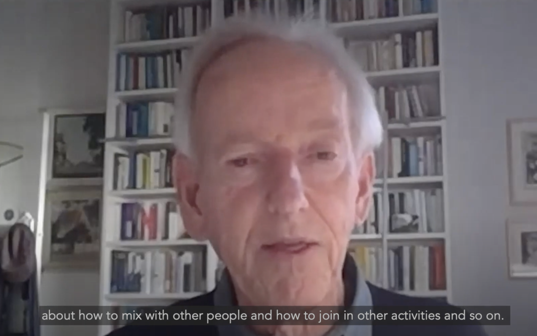An elderly man stands in front of a wall of books & addresses the camera