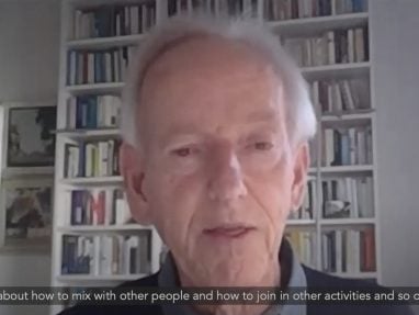 An elderly man stands in front of a wall of books & addresses the camera