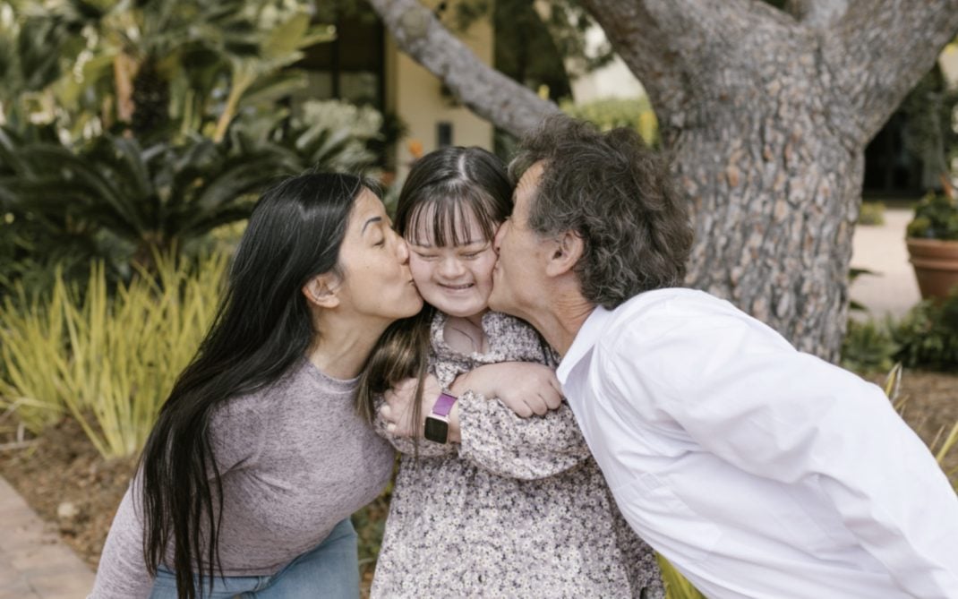 A dark haired woman & dark haired man, leaning in to playfully kiss either cheek of their dark haired child