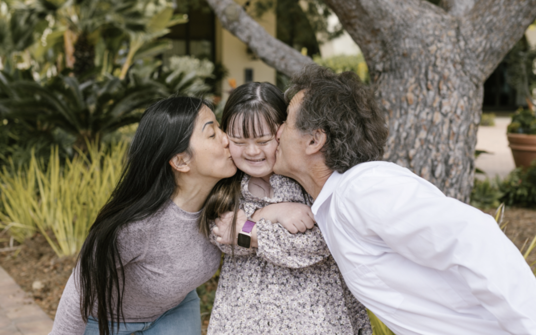 A dark haired woman & dark haired man, leaning in to playfully kiss either cheek of their dark haired child