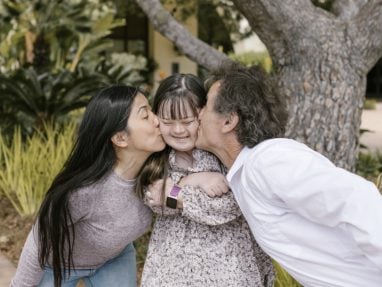 A dark haired woman & dark haired man, leaning in to playfully kiss either cheek of their dark haired child