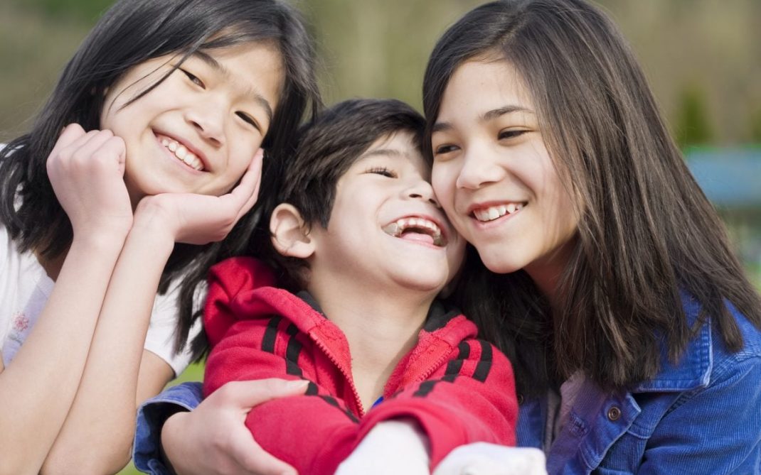 Three smiling children, two girls and one boy, pose closely together outdoors. The boy in the center wears a red jacket, while the girls on either side wear blue and white. All look happy and are enjoying each others company.