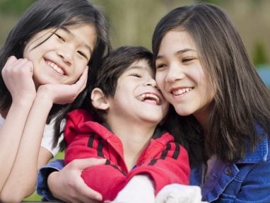 Three smiling children, two girls and one boy, pose closely together outdoors. The boy in the center wears a red jacket, while the girls on either side wear blue and white. All look happy and are enjoying each others company.