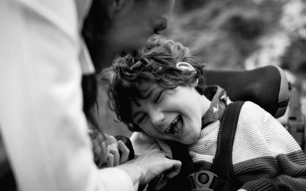 A child in a wheelchair laughs joyfully as an adult beside them smiles and gently touches their arm. The photo is in black and white, capturing a moment of happiness and connection.