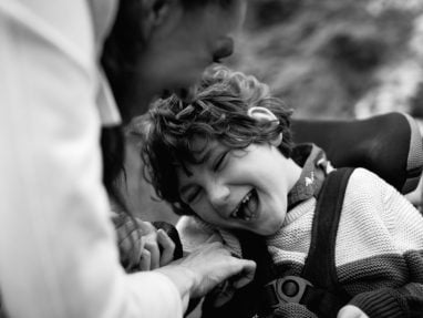 A child in a wheelchair laughs joyfully as an adult beside them smiles and gently touches their arm. The photo is in black and white, capturing a moment of happiness and connection.