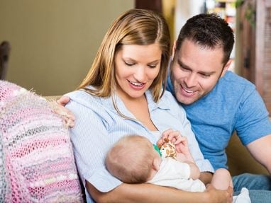 A smiling woman and man sit on a couch, looking lovingly at a baby in the womans arms. The baby holds a small toy, and a pink crocheted blanket is draped over the back of the couch.