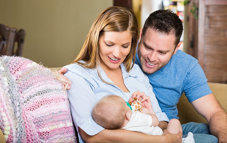 A smiling woman and man sit on a couch, looking lovingly at a baby in the womans arms. The baby holds a small toy, and a pink crocheted blanket is draped over the back of the couch.