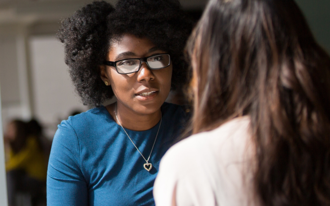 A dark skinned woman, sits at a table consoling another woman. Her hands are folded together & she shows engagement in their conversation.