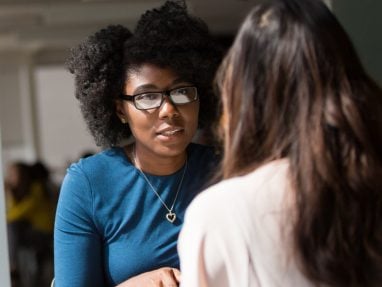 A dark skinned woman, sits at a table consoling another woman. Her hands are folded together & she shows engagement in their conversation.