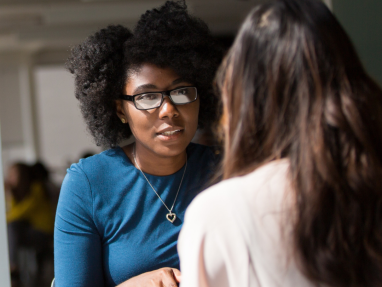 A dark skinned woman, sits at a table consoling another woman. Her hands are folded together & she shows engagement in their conversation.