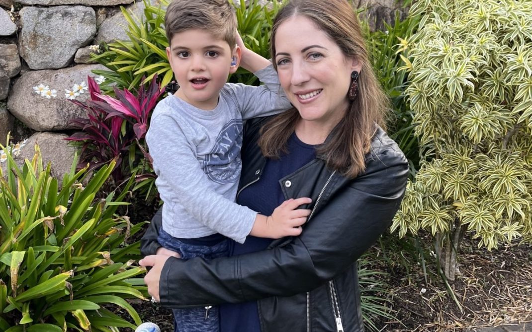 A woman in a black jacket smiles while holding a young boy wearing a gray shirt and blue shoes. They are outdoors, surrounded by green plants, colorful flowers, and a stone wall in the background.