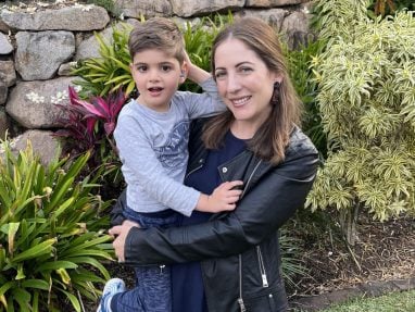 A woman in a black jacket smiles while holding a young boy wearing a gray shirt and blue shoes. They are outdoors, surrounded by green plants, colorful flowers, and a stone wall in the background.