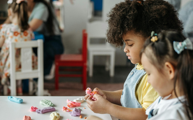 Two young children sit at a table shaping colorful modeling clay, while other kids and adults interact at tables in the background in a bright classroom or daycare setting.