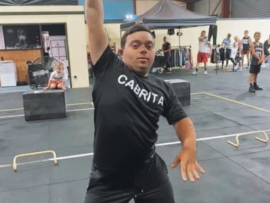 A young man wearing a black CABRITA shirt lifts a dumbbell overhead in a gym. People and gym equipment are visible in the background.