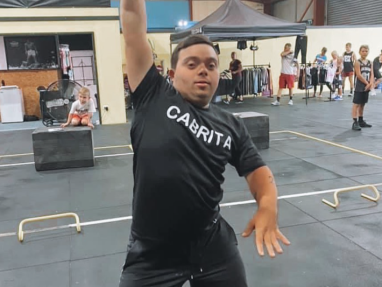 A young man wearing a black CABRITA shirt lifts a dumbbell overhead in a gym. People and gym equipment are visible in the background.