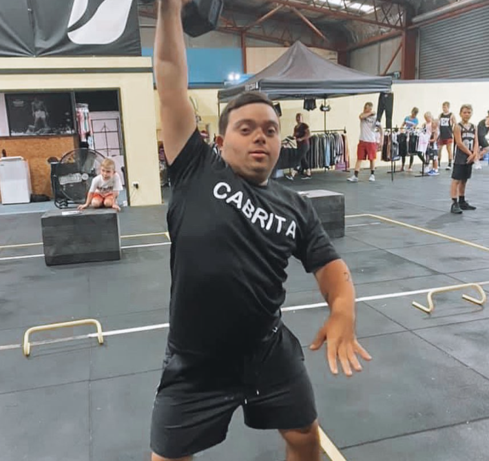A young man wearing a black CABRITA shirt lifts a dumbbell overhead in a gym. People and gym equipment are visible in the background.