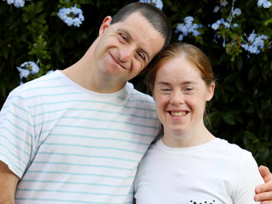 A man and woman, both smiling and wearing white shirts, stand close together with the man’s arm around the woman’s shoulders. Blue flowers and green foliage fill the background.