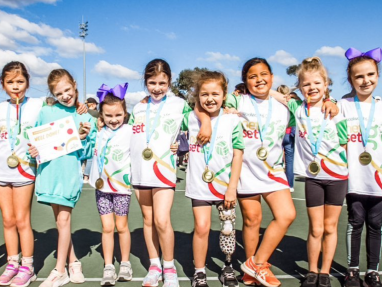 A group of eight young girls wearing medals and white sports shirts stand together, smiling with arms around each other on an outdoor court under a blue sky with clouds.