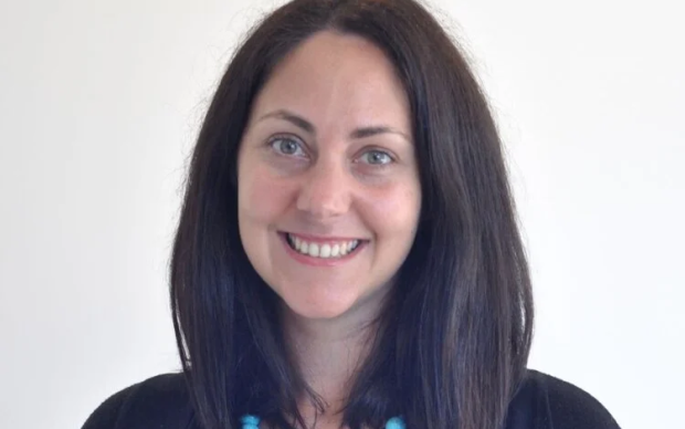 A woman with straight, shoulder-length dark hair smiles at the camera. She is wearing a black top, a black cardigan, and a chunky turquoise bead necklace. The background is plain white.