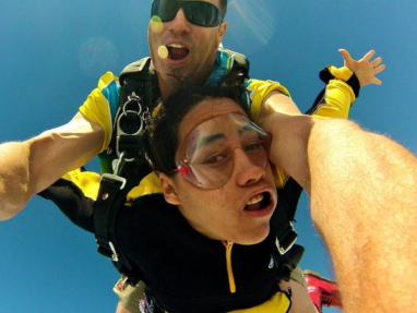 Two people are skydiving in tandem against a clear blue sky, both wearing yellow and black jumpsuits. The instructor is behind, holding the person in front, who looks nervous, while the instructor is smiling.