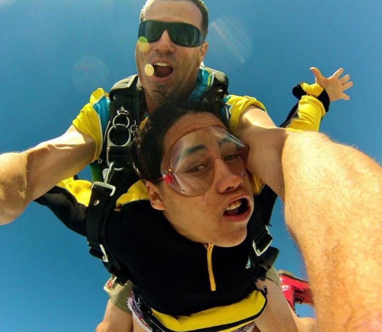 Two people are skydiving in tandem against a clear blue sky, both wearing yellow and black jumpsuits. The instructor is behind, holding the person in front, who looks nervous, while the instructor is smiling.