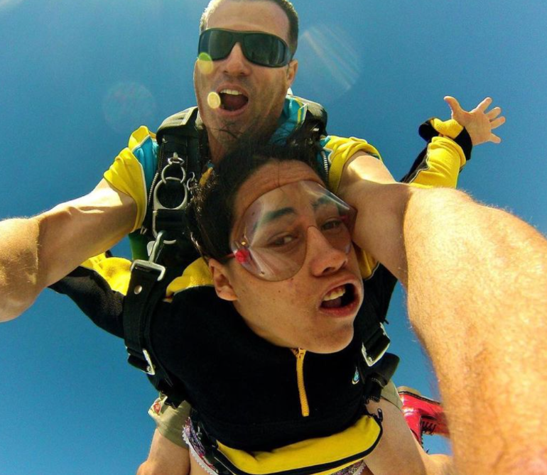 Two people are skydiving in tandem against a clear blue sky, both wearing yellow and black jumpsuits. The instructor is behind, holding the person in front, who looks nervous, while the instructor is smiling.