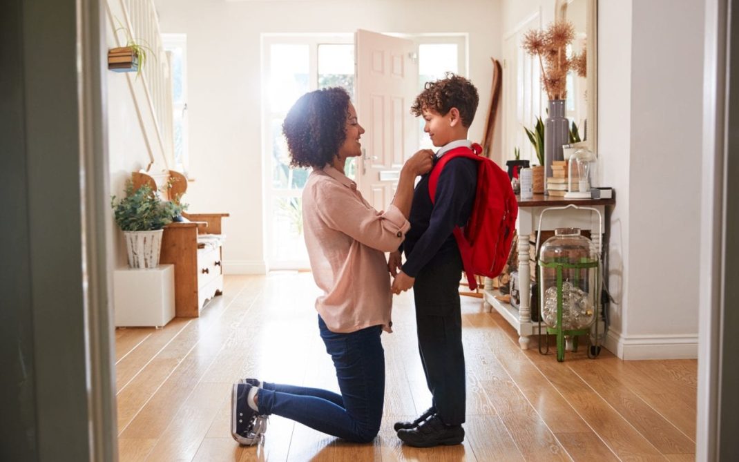 A woman kneels to adjust a young boy’s red backpack in a bright hallway. The boy, dressed in a school uniform, smiles at her as sunlight streams through the open front door behind them.