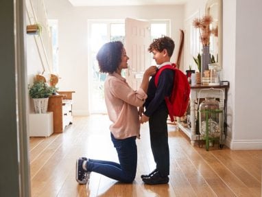 A woman kneels to adjust a young boy’s red backpack in a bright hallway. The boy, dressed in a school uniform, smiles at her as sunlight streams through the open front door behind them.