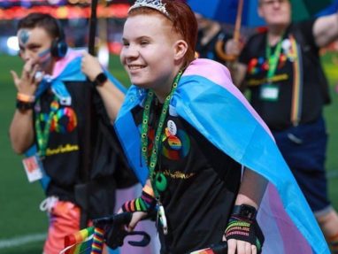 A smiling person wearing a tiara and a transgender pride flag as a cape holds rainbow flags and uses crutches at a colorful outdoor event, with people and rainbow umbrellas in the background.