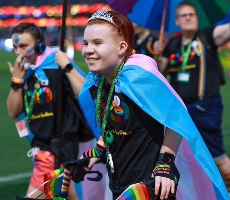 A smiling person wearing a tiara and a transgender pride flag as a cape holds rainbow flags and uses crutches at a colorful outdoor event, with people and rainbow umbrellas in the background.