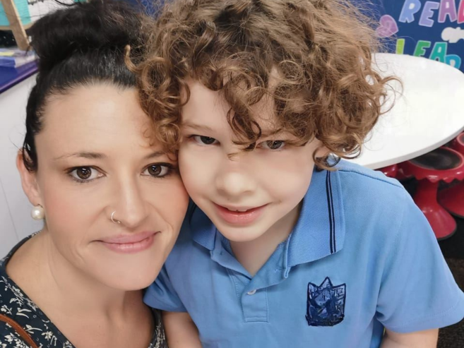 A woman with dark hair in a bun and a young boy with curly hair and a blue shirt smile closely together, posing for a selfie in a colorful room with a round table and red chairs in the background.