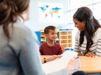 A young boy sits between two women at a table in a classroom, smiling and looking at one of them. One woman holds a notepad and pen. A world map is visible on the wall in the background.