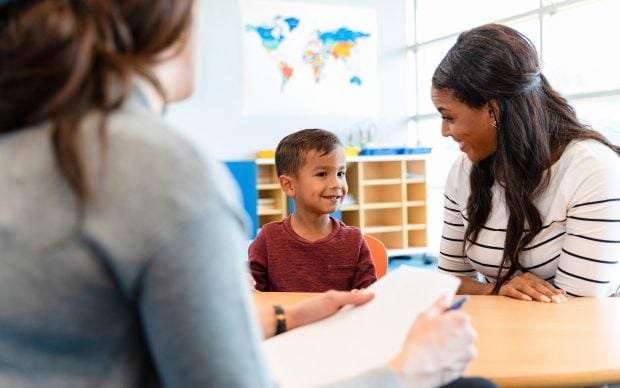 A young boy sits between two women at a table in a classroom, smiling and looking at one of them. One woman holds a notepad and pen. A world map is visible on the wall in the background.
