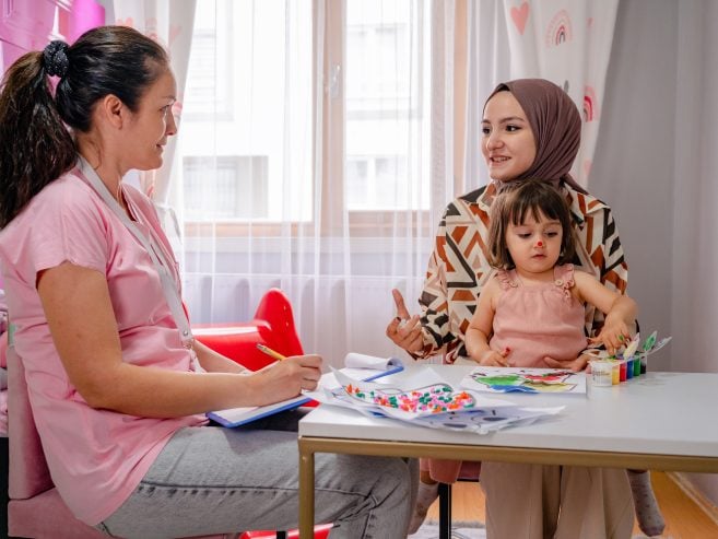 A woman in a hijab sits with her young daughter at a small table, talking to a preschool educator who is taking notes. Art supplies and colorful drawings are on the table in a bright room.