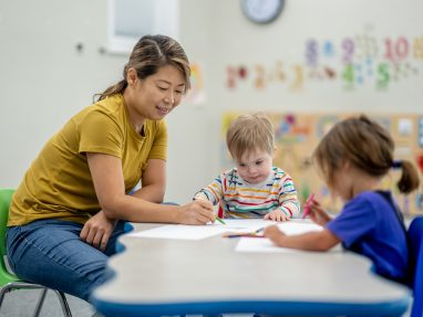A woman helps two young children with drawing at a classroom table. The children are focused on their papers, and colorful numbers decorate the wall in the background.