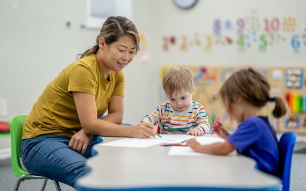 A woman helps two young children with drawing at a classroom table. The children are focused on their papers, and colorful numbers decorate the wall in the background.