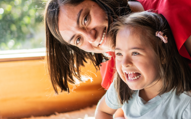 A woman smiles and hugs a young girl with Down syndrome. They are sitting by a sunny window, both looking happy and joyful.