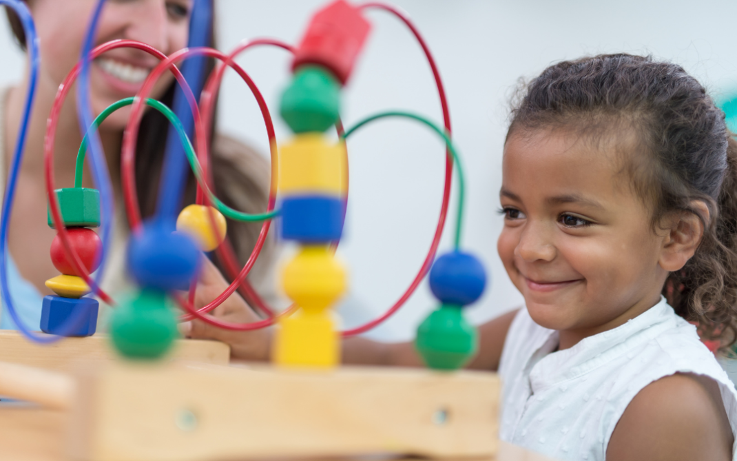 A young child smiles while playing with a bead maze toy, sitting at a table. An adult in the background also smiles, watching and engaging with the child in a bright, classroom-like setting.