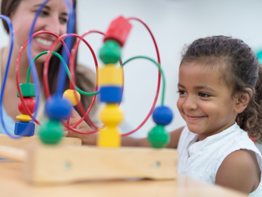 A young child smiles while playing with a bead maze toy, sitting at a table. An adult in the background also smiles, watching and engaging with the child in a bright, classroom-like setting.