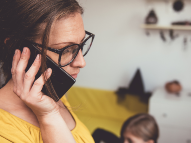 A woman wearing glasses and a yellow top talks on her smartphone, looking concerned. In the blurred background, a child sits on the floor and Halloween decorations are visible on the wall and shelf.