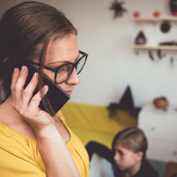 A woman wearing glasses and a yellow top talks on her smartphone, looking concerned. In the blurred background, a child sits on the floor and Halloween decorations are visible on the wall and shelf.