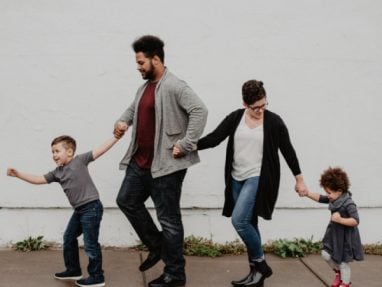 A family of four, two adults and two children, hold hands and walk in a line along a sidewalk in front of a white wall, smiling and playfully stepping together.